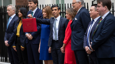 Politicians stand outside downing street, Rishi Sunak holds a red suitcase up