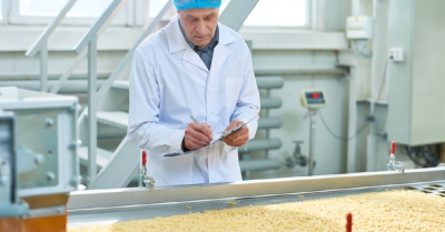 A man with blue hair covering and white coat writes something on a board. He is in a factory with yellow chips on a conveyor belt