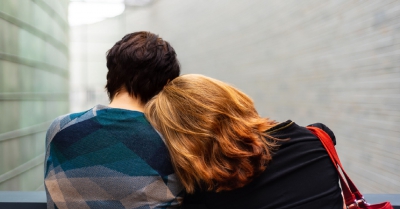 two people one with short black hair and one with medium length red hair sit together, the person with red hair rests their head on the other persons shoulder