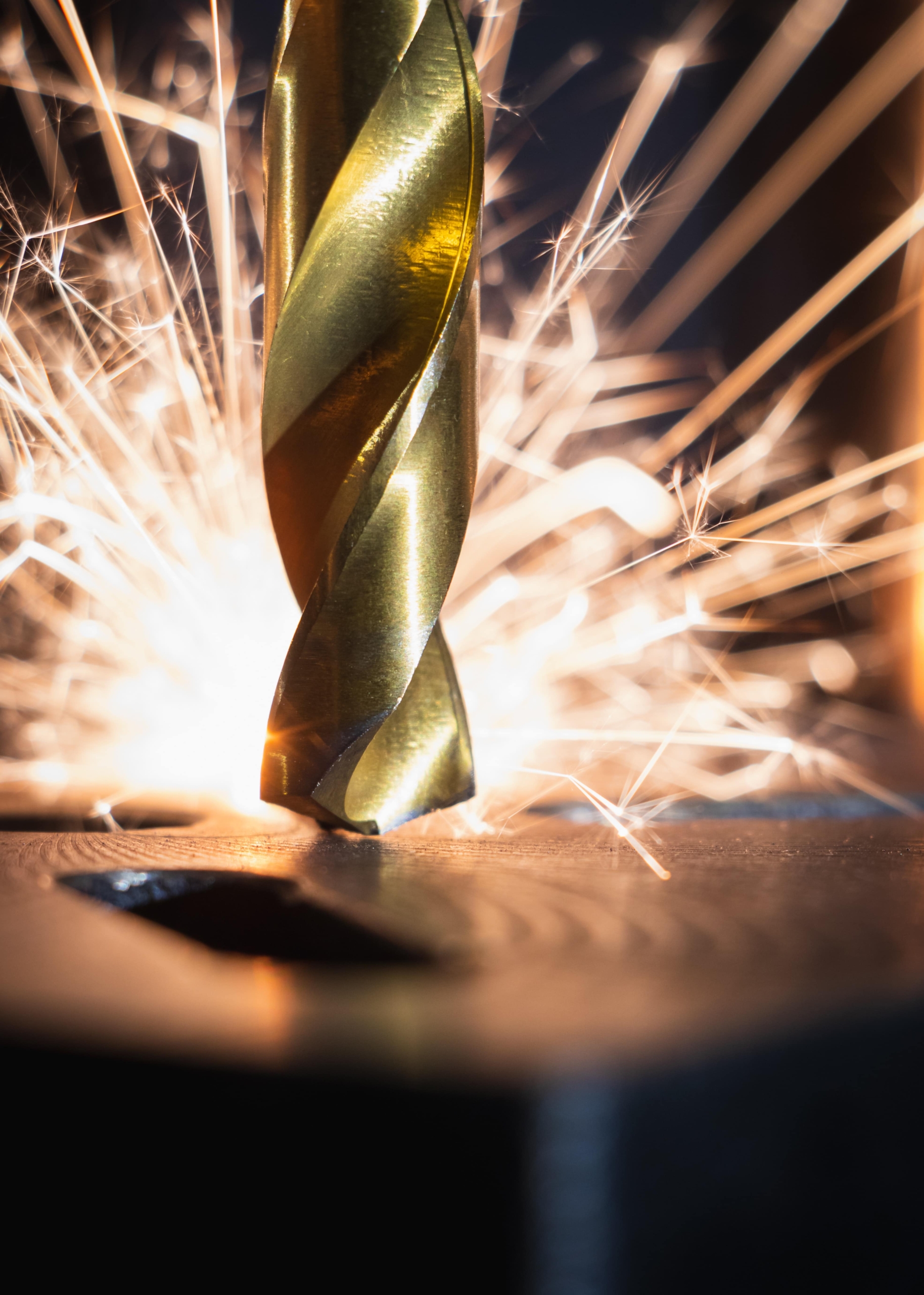 A gold spiral drill bit touches a metal board and sparks fly around it