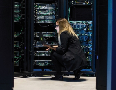 A lady sqats down next to servers with her laptop in her hand, she is looking at the screen